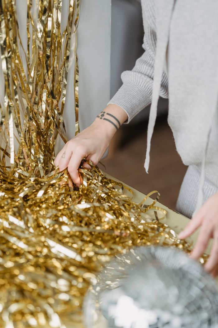 Crop unrecognizable tattooed female touching pile of shiny tinsel at table in light house